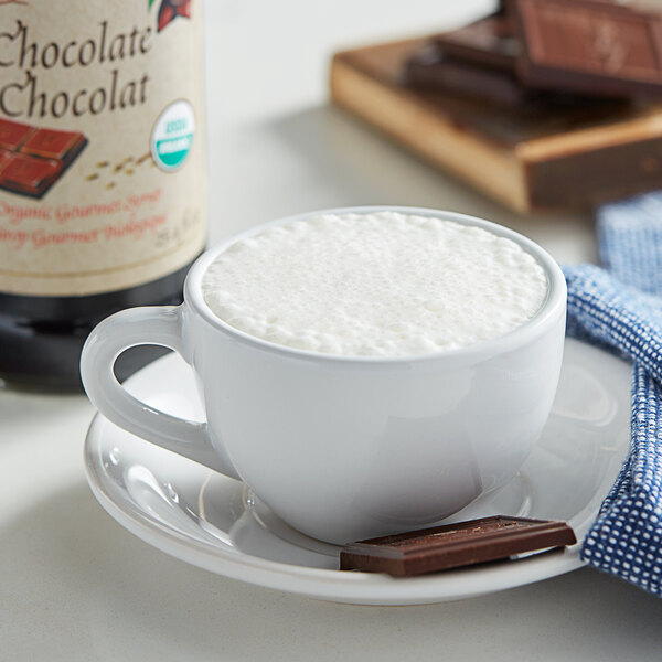 A white cup of hot chocolate with foam on top and a bottle of Monin Organic Chocolate Flavoring.