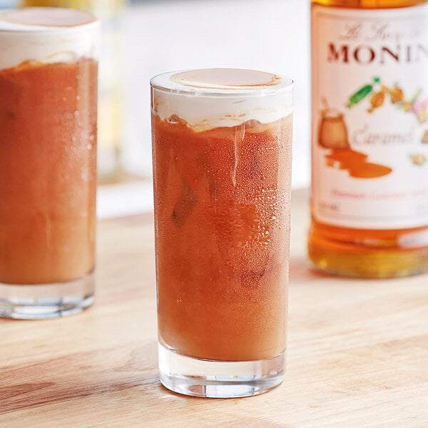 A glass of brown liquid on a table with a bottle of Monin Premium Caramel Flavoring Syrup.