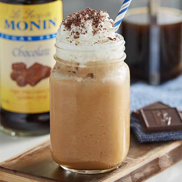 A glass of brown liquid with whipped cream and chocolate chips next to a bottle of Monin Sugar Free Chocolate Flavoring Syrup.