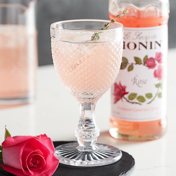 A wine glass filled with pink liquid next to a bottle of Monin Premium Rose Flavoring Syrup.