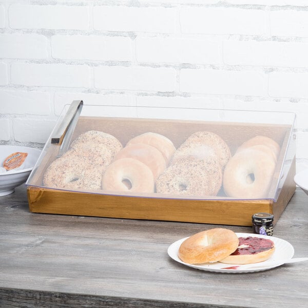 A Cal-Mil Madera display case with a clear lid on a counter with a plate of bagels inside.