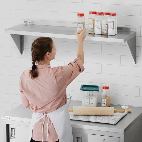 A woman in a white shirt standing on a Regency stainless steel wall shelf in a professional kitchen holding a container of spices.