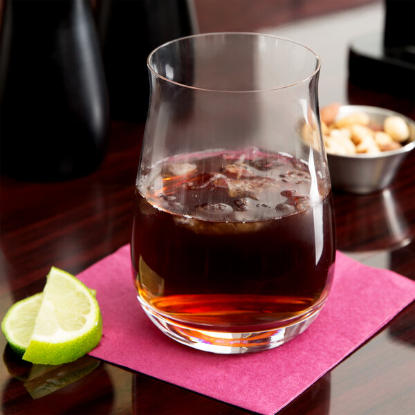 A Spiegelau whiskey tumbler filled with brown liquid and ice with a lime wedge on the table.