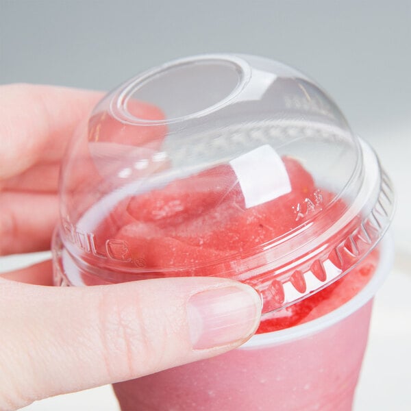 A hand holding a Dart clear plastic cup with a pink drink inside and a clear plastic dome lid with a hole in it.
