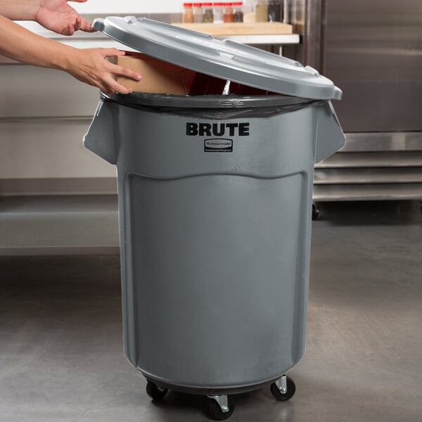 A person's hand opening the lid of a grey Rubbermaid commercial trash can.