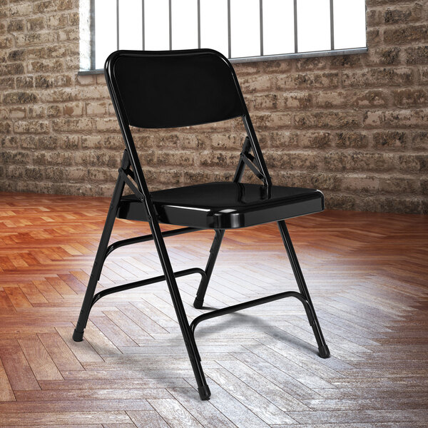 A black National Public Seating folding chair in front of a brick wall.