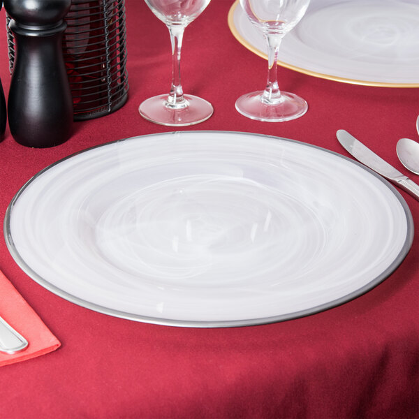 A white Charge It by Jay alabaster glass charger plate with silver rim on a table with silverware and wine glasses.