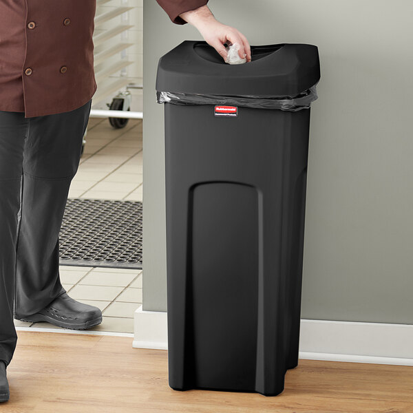 A man in a chef's uniform standing next to a Rubbermaid black square commercial trash can.