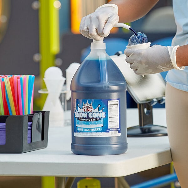 A person in gloves pouring Carnival King Blue Raspberry syrup from a large blue container into a plastic container.