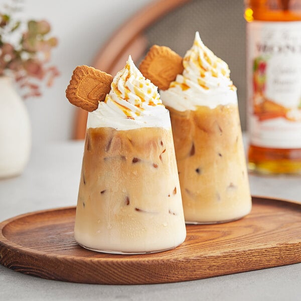 Two glasses of iced coffee with whipped cream and cookies on a table with a bottle of Monin Cookie Butter Flavoring Syrup.
