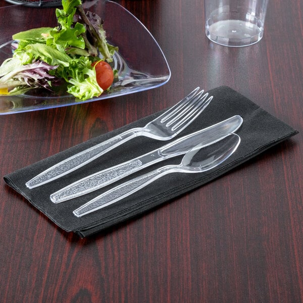 A Visions black pre-rolled linen-feel napkin with clear heavy weight plastic fork and knife on a table next to a salad