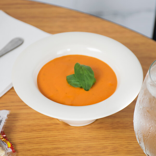 A bowl of orange soup with basil leaves on top in a GET Ivory Melamine bowl on a table.