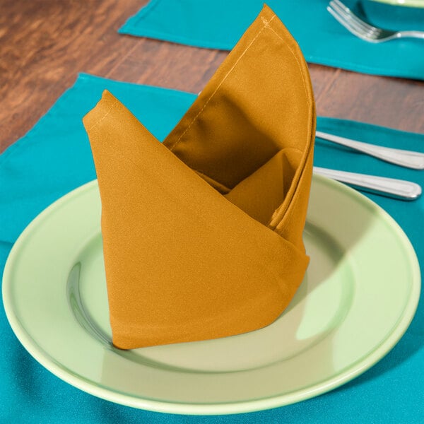 A folded Intedge gold polycotton napkin on a plate with silverware.