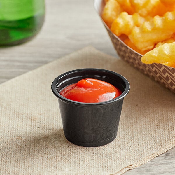 A black plastic souffle cup filled with french fries on a table with a bowl of ketchup.