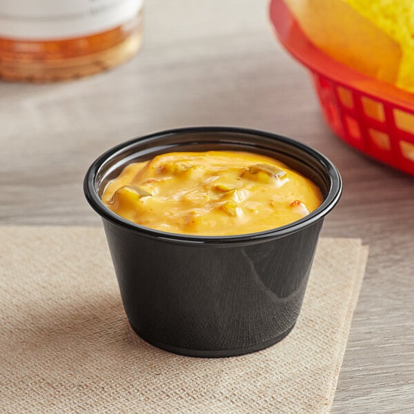 A black plastic souffle cup of yellow sauce on a table with a bowl of chips.