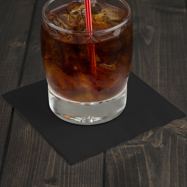 A glass of ice and liquid with a black Creative Converting beverage napkin on a table.