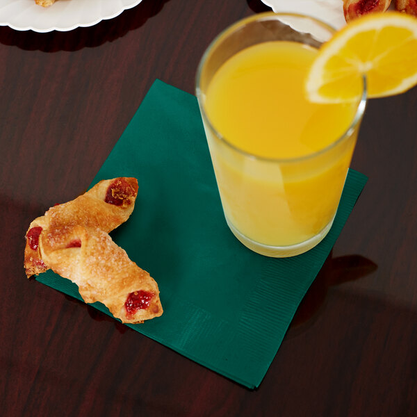 A table with a glass of orange juice and a pack of Creative Converting Hunter Green Beverage Napkins on it.