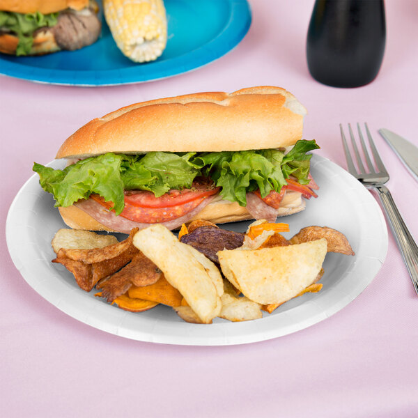 A sandwich with lettuce and tomato on a white paper plate with potato chips and corn.