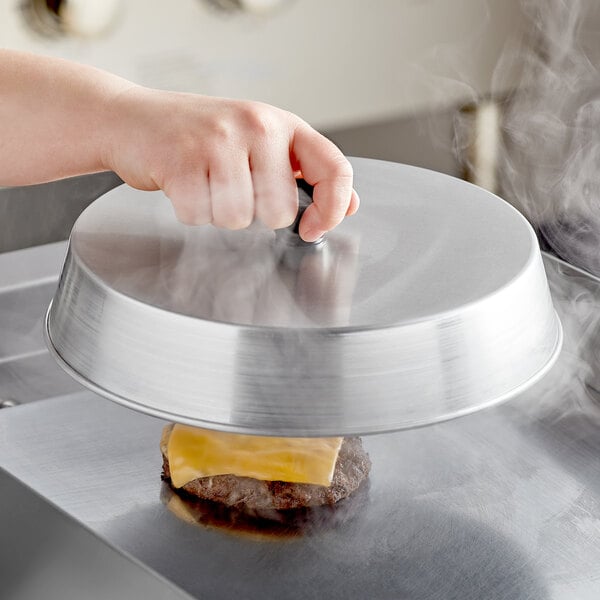 A person holding an American Metalcraft aluminum basting cover over a burger on a grill.