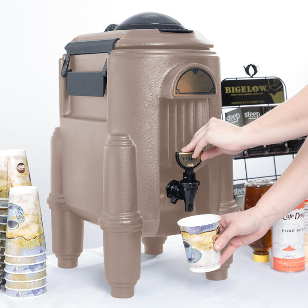 A person's hand pressing a Cambro dark taupe insulated beverage dispenser on a counter to pour a drink.