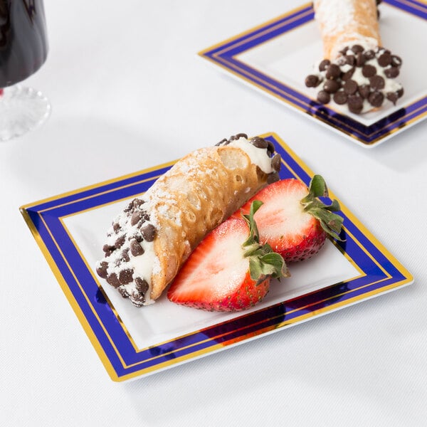 A Fineline white plastic square plate with blue rim and gold bands holding a cannoli and strawberries.