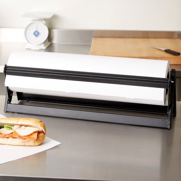 A Bulman black paper dispenser with a serrated blade holding a roll of paper on a black counter next to a sandwich.