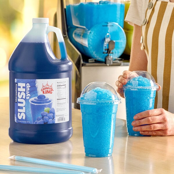 A gallon jug of Carnival King blue raspberry slush syrup concentrate next to two cups of blue slushy drinks.