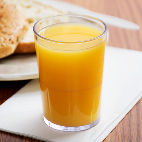A Cambro clear plastic tumbler filled with orange juice on a table with bread.