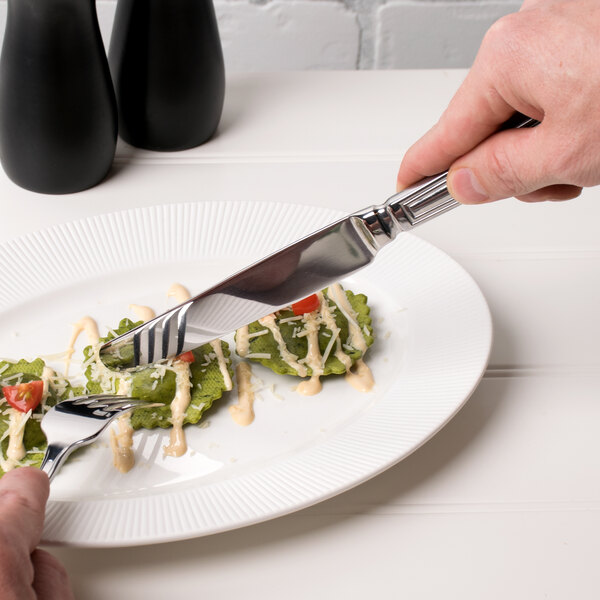 A person holding a Libbey Fairfield stainless steel dinner knife and fork over a plate of food.