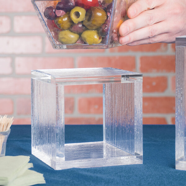 A hand holding a bowl of olives over an American Metalcraft clear square acrylic riser.