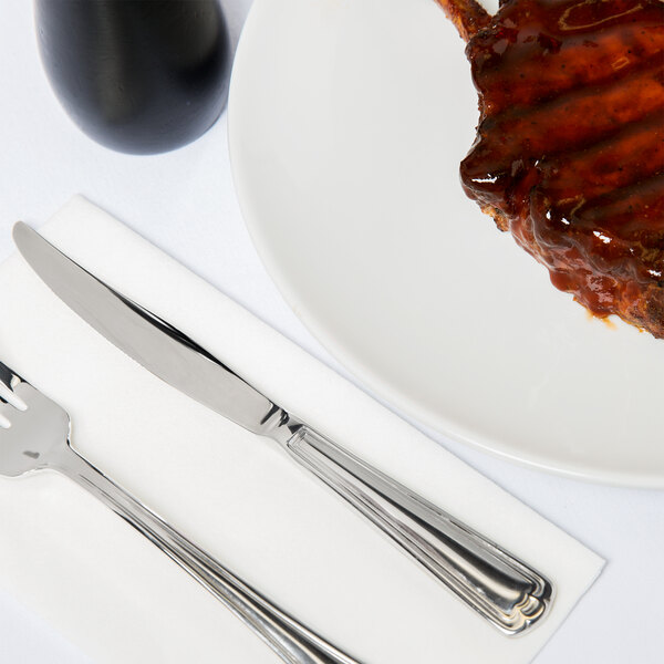 A white plate with a steak and sauce next to a Libbey Fairfield stainless steel steak knife and fork.