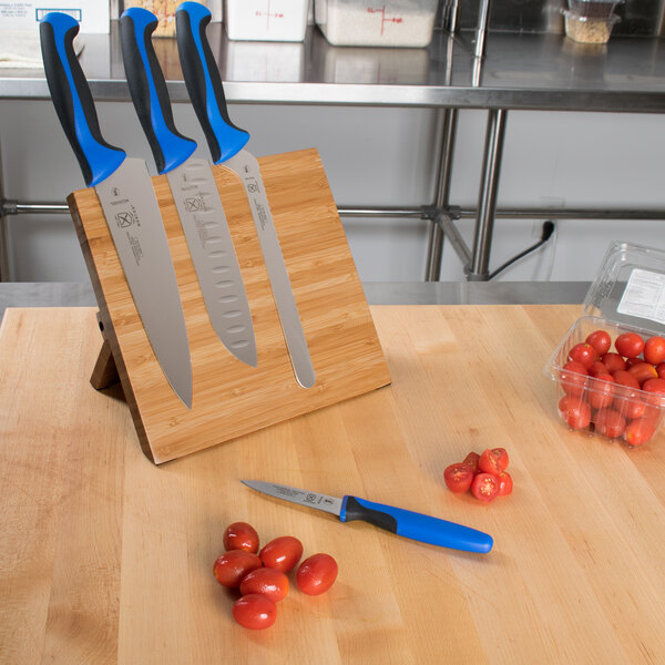 A Mercer Culinary bamboo magnetic board with a group of blue-handled knives on it next to tomatoes.