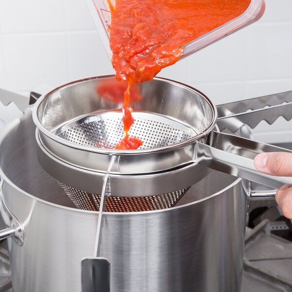 A person using a Vollrath Coarse China Cap Strainer to pour red sauce into a pot.