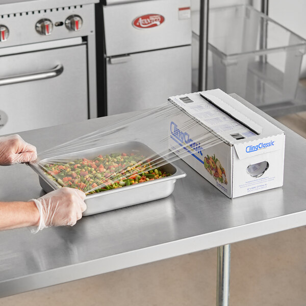 A person in gloves using a serrated cutter to dispense Berry standard plastic film over a tray of food.