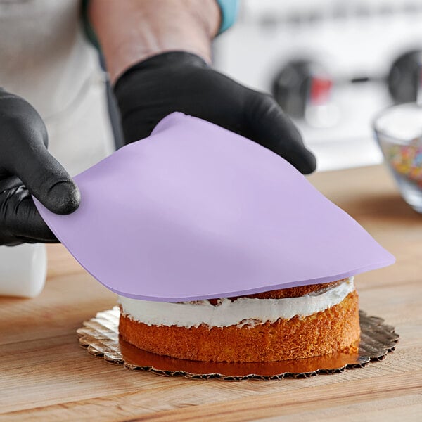 A person wearing black gloves and using a purple spatula to cut a cake covered in Satin Ice Lavender Vanilla rolled fondant.