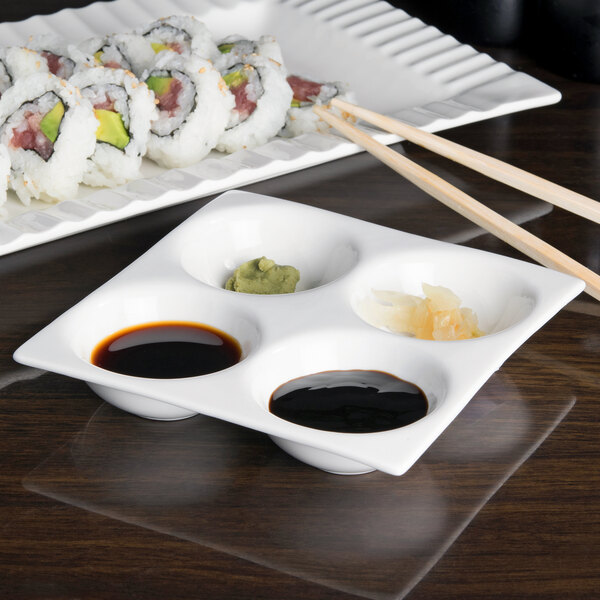 An Arcoroc white porcelain dish with four compartments filled with sushi, soy sauce, and sauces.