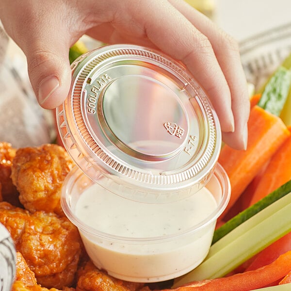 A hand holding a plastic container with a clear lid over white sauce on a plate of food.