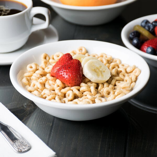 A close-up of a Libbey Aluma White porcelain cereal bowl filled with cereal, strawberries, and bananas on a table with a cup of coffee.