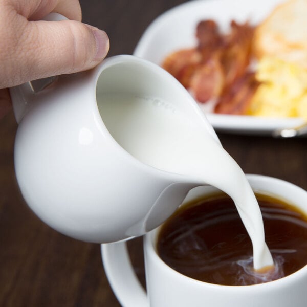 A person pouring milk into a cup of coffee using a Libbey white porcelain creamer.