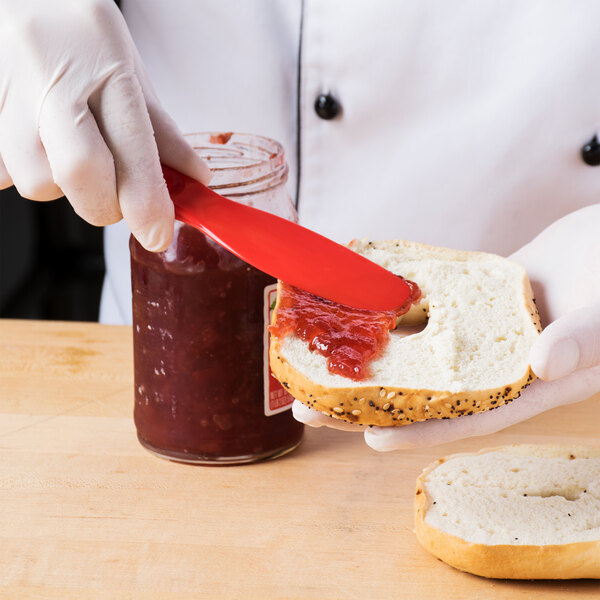 A gloved hand uses a red HS Inc. sandwich spreader to spread jam on a bagel.