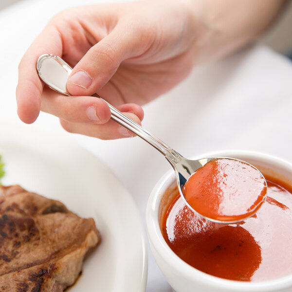 A person using a Reserve by Libbey stainless steel bouillon spoon to eat red sauce over a bowl.
