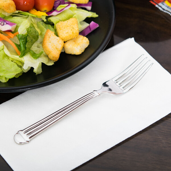 A Libbey Aegean stainless steel salad fork on a napkin next to a plate of salad.
