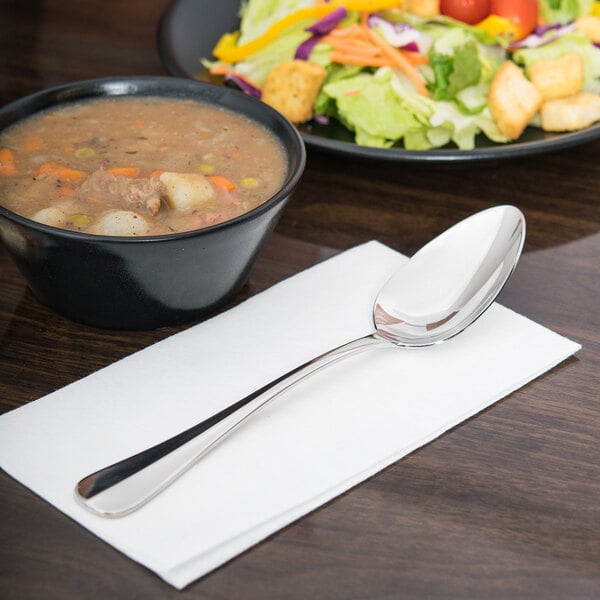 A Libbey Baguette II stainless steel serving spoon on a napkin next to a bowl of soup.