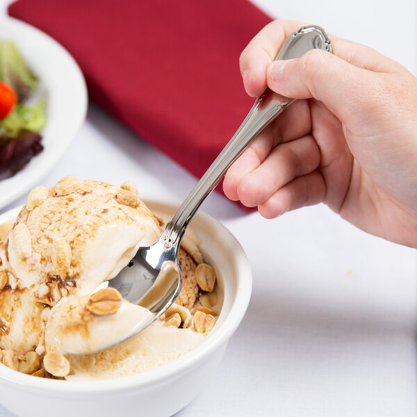 A hand holding a Reserve by Libbey Baroque stainless steel dessert spoon over a bowl of ice cream.