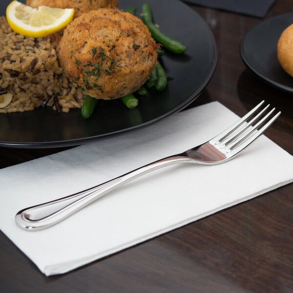 A Libbey stainless steel dinner fork on a napkin next to a plate of food with a lemon wedge.