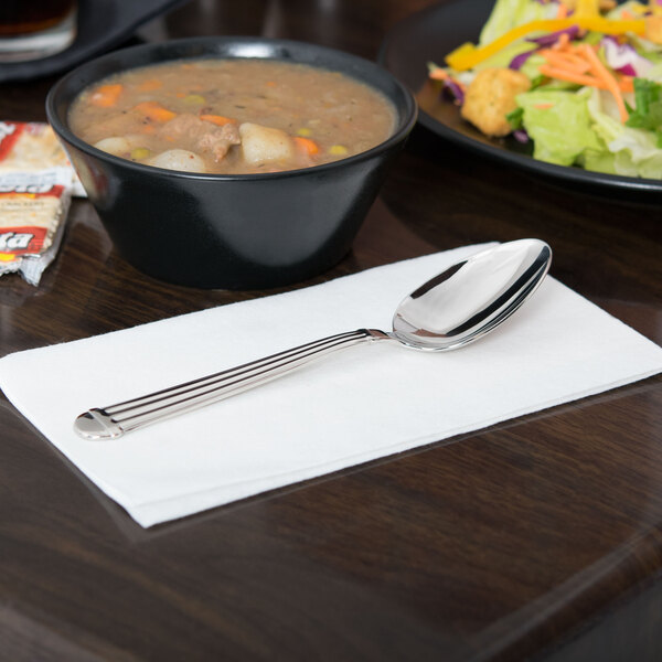 A Libbey extra heavy weight stainless steel dessert spoon on a white napkin next to a bowl of soup, salad, and crackers.
