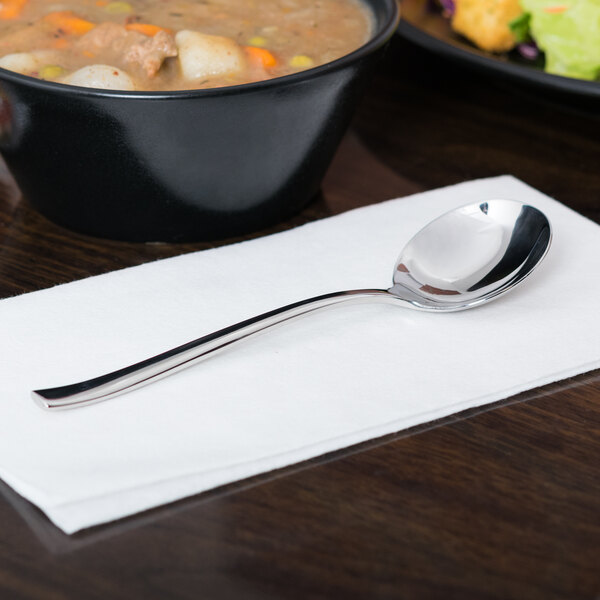 A Libbey stainless steel bouillon spoon on a napkin next to a bowl of soup.