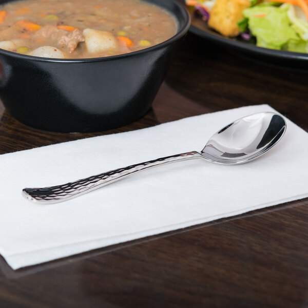 A stainless steel Libbey bouillon spoon on a white napkin next to a bowl of soup.