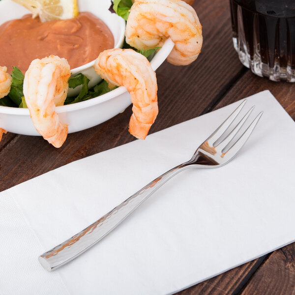 A Libbey stainless steel cocktail fork on a napkin next to a bowl of shrimp.