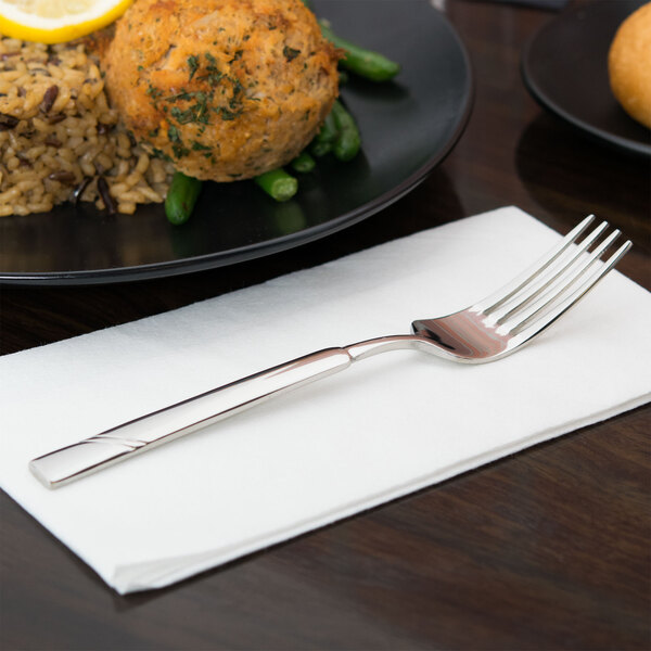 A Libbey stainless steel dessert fork on a white napkin next to a plate of food.